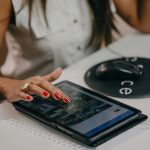 A woman with manicured nails uses a tablet on a desk, technology in focus.