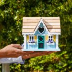 Close-up of a man's hands holding a decorative miniature house against foliage backdrop.